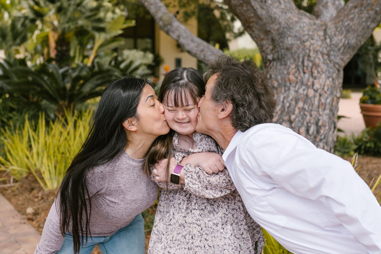 A young woman with Down syndrome laughs as her parents kiss her cheeks in a leafy garden.