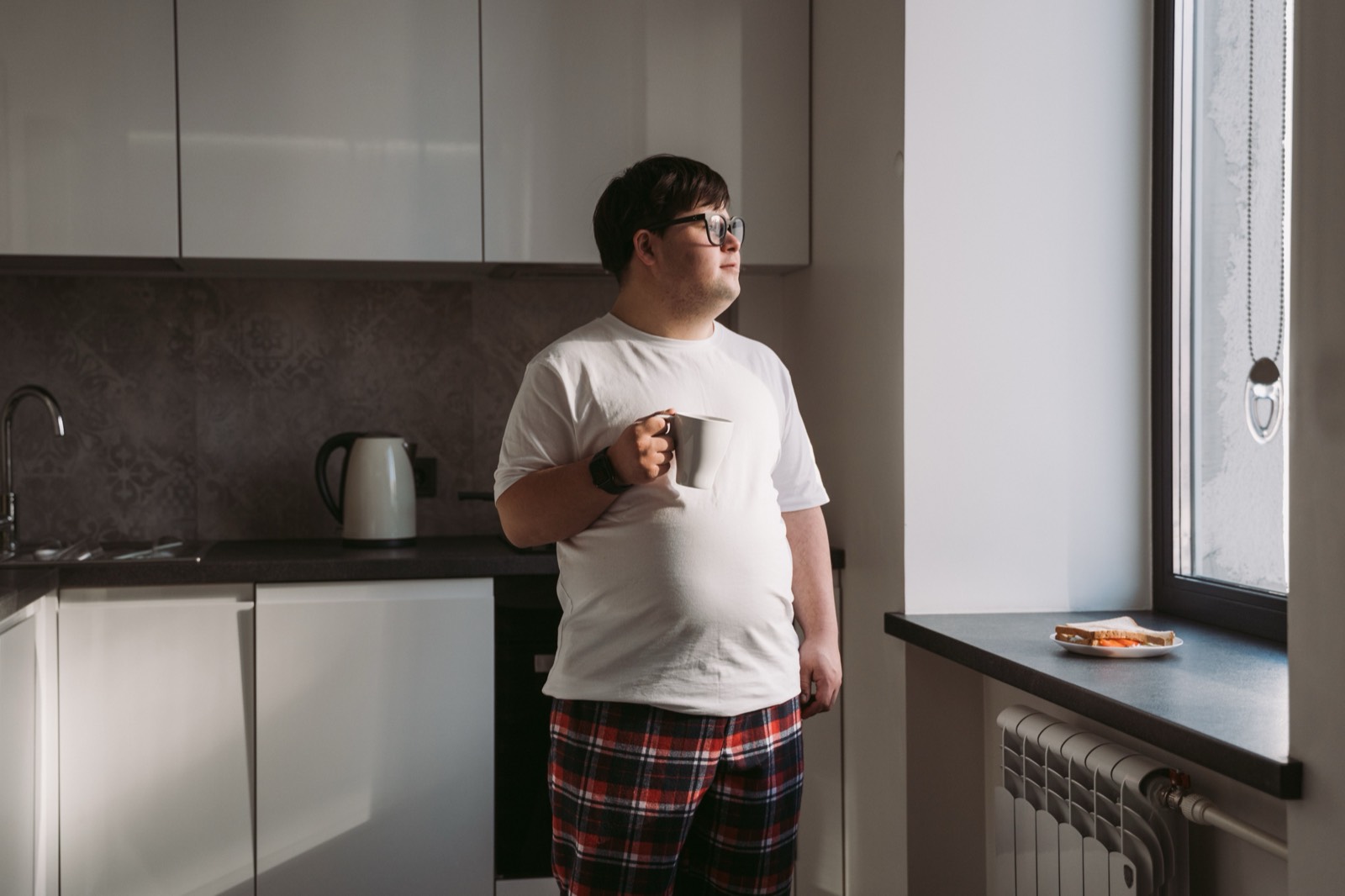 A young man with Down syndrome holds a mug of coffee and looks out the window of his kitchen in morning light.