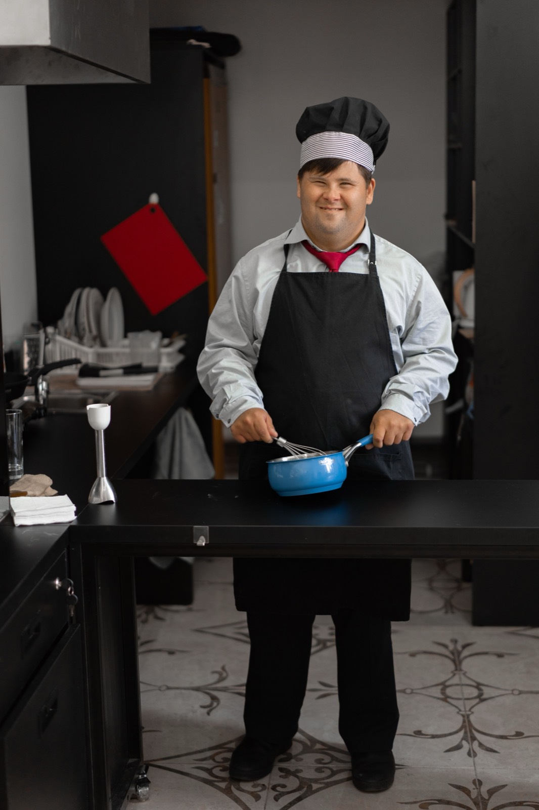 A man with Down syndrome in an apron and chef's hat smiles while whisking in a saucepan in a kitchen.